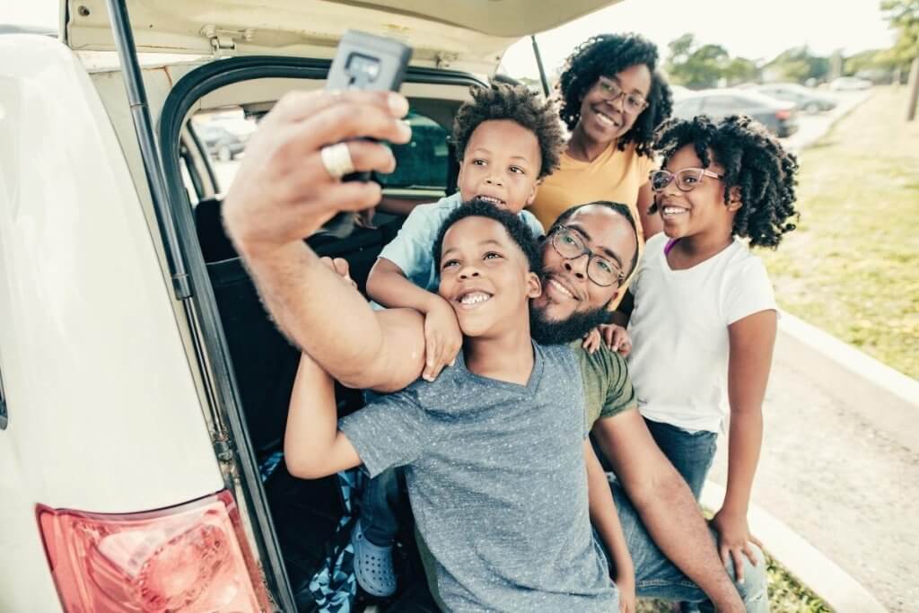 a happy family in the back of their car smiling and posing for a cellphone photo